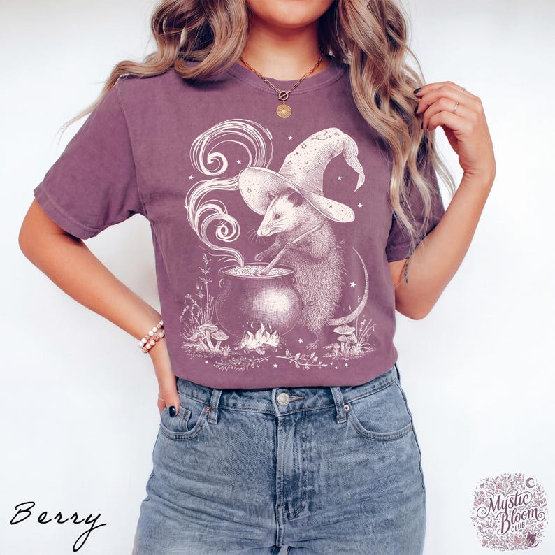 May include: A berry-colored short-sleeve t-shirt featuring a white graphic print of an opossum wearing a wizard hat and stirring a bubbling cauldron. The design includes swirling smoke, small mushrooms, and stars, set against a solid fabric.
