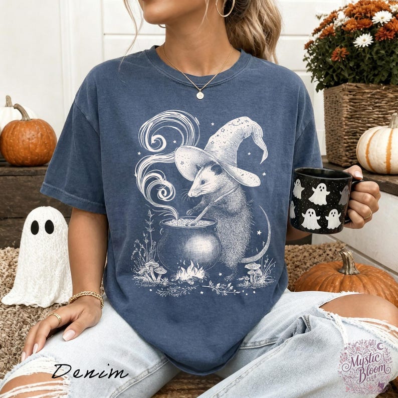 May include: A blue short-sleeve t-shirt featuring a white graphic print of an opossum wearing a witch hat and stirring a cauldron. The design includes swirling smoke, small mushrooms, and stars. The shirt has a crew neckline and a relaxed fit.