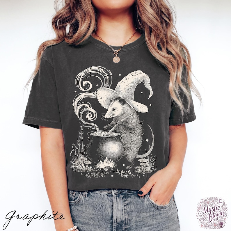May include: A charcoal gray short-sleeve t-shirt featuring a white graphic illustration of an opossum wearing a wizard hat and stirring a bubbling cauldron over a small fire, surrounded by swirling smoke, stars, and small mushrooms.