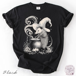 May include: A black short-sleeved t-shirt featuring a white graphic illustration of an opossum wearing a star-patterned wizard hat. The opossum is stirring a steaming cauldron over a small fire, surrounded by mushrooms and small stars.