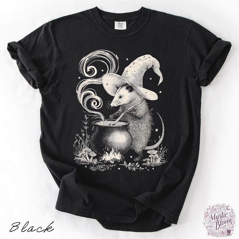 May include: A black short-sleeved t-shirt featuring a white graphic illustration of an opossum wearing a star-patterned wizard hat. The opossum is stirring a steaming cauldron over a small fire, surrounded by mushrooms and small stars.