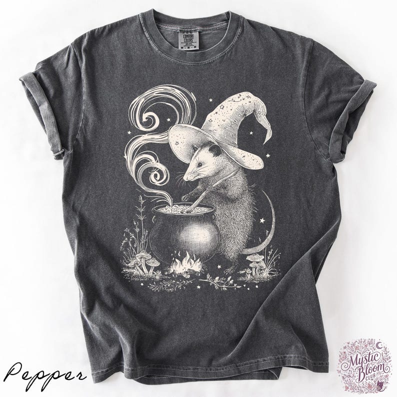 May include: A charcoal gray short-sleeve t-shirt featuring a white graphic illustration of an opossum wearing a wizard hat and stirring a bubbling cauldron over a small fire, surrounded by swirling smoke, stars, and small mushrooms.
