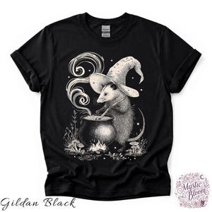 May include: A black t-shirt featuring a white graphic illustration of an opossum wearing a wizard hat and stirring a steaming cauldron over a small fire. The design includes small mushrooms, stars, and foliage surrounding the base of the cauldron.