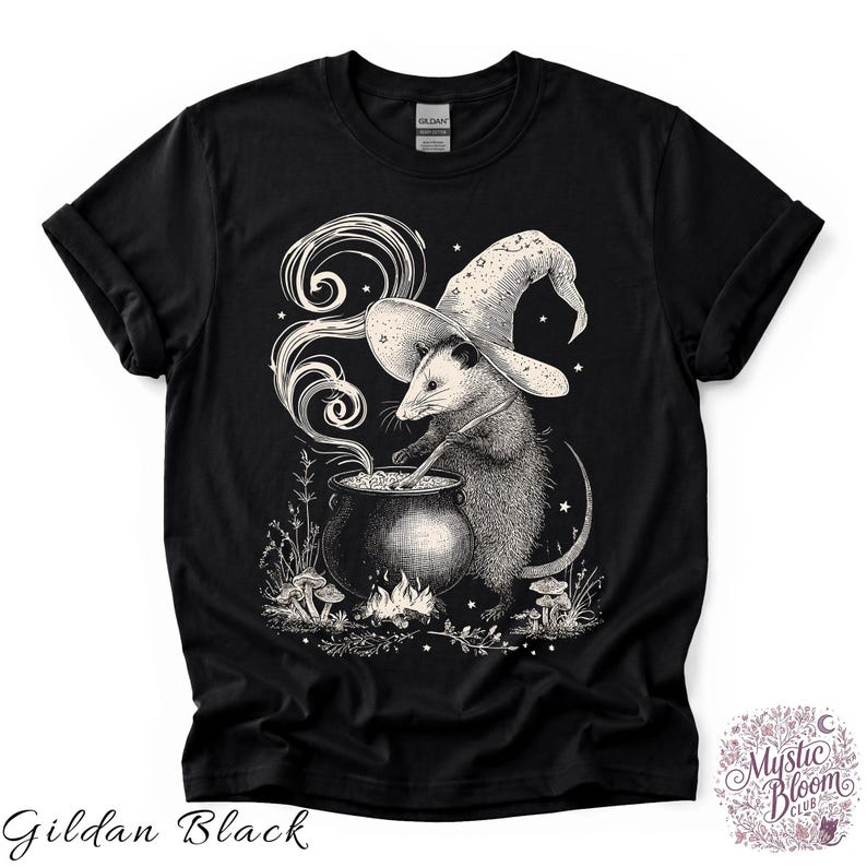 May include: A black t-shirt featuring a white graphic illustration of an opossum wearing a wizard hat and stirring a steaming cauldron over a small fire. The design includes small mushrooms, stars, and foliage surrounding the base of the cauldron.