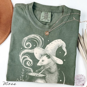 May include: A moss green short-sleeve t-shirt featuring a graphic of an opossum wearing a wizard hat and stirring a cauldron with swirling smoke. The shirt has a Comfort Colors label at the neck and a thin gold-toned chain necklace resting on it.