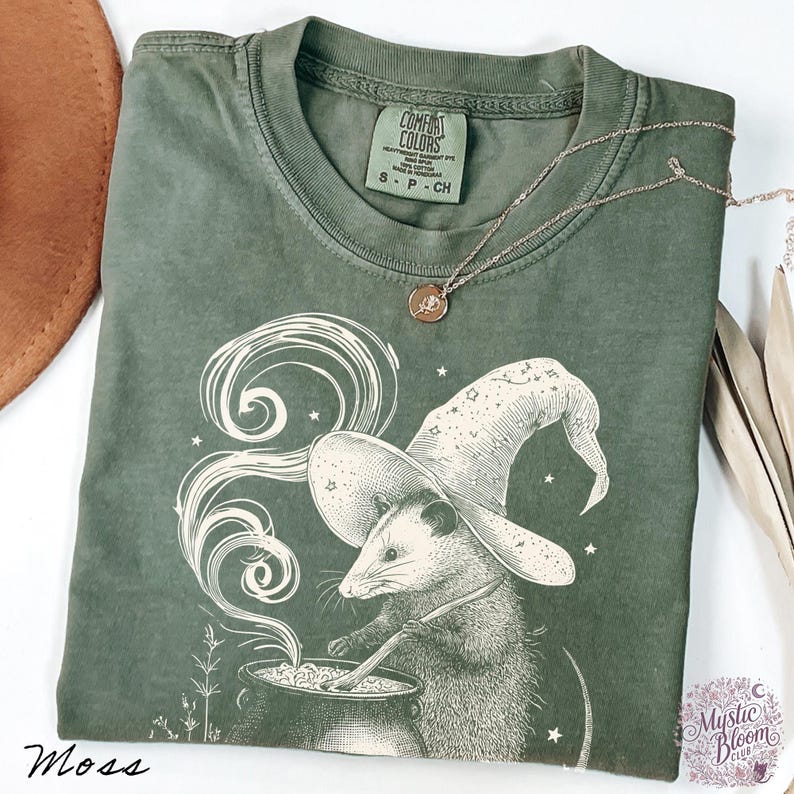 May include: A moss green short-sleeve t-shirt featuring a graphic of an opossum wearing a wizard hat and stirring a cauldron with swirling smoke. The shirt has a Comfort Colors label at the neck and a thin gold-toned chain necklace resting on it.