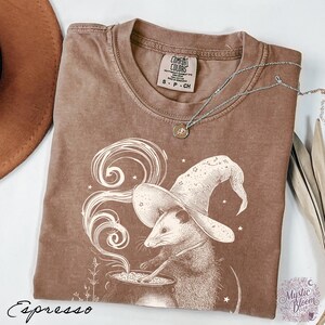 May include: A brown short-sleeved t-shirt featuring a graphic of an opossum wearing a wizard hat and stirring a cauldron with swirling smoke. The shirt has a crew neckline and a Comfort Colors brand label. The word Espresso is printed below.