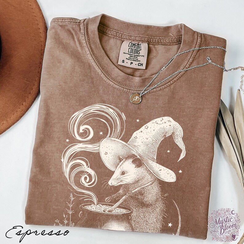 May include: A brown short-sleeved t-shirt featuring a graphic of an opossum wearing a wizard hat and stirring a cauldron with swirling smoke. The shirt has a crew neckline and a Comfort Colors brand label. The word Espresso is printed below.