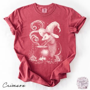 May include: A crimson short-sleeve t-shirt featuring a white graphic illustration of an opossum wearing a wizard hat and stirring a cauldron over a small fire. The design includes swirling smoke, stars, and mushrooms, with the text Comfort Colors.