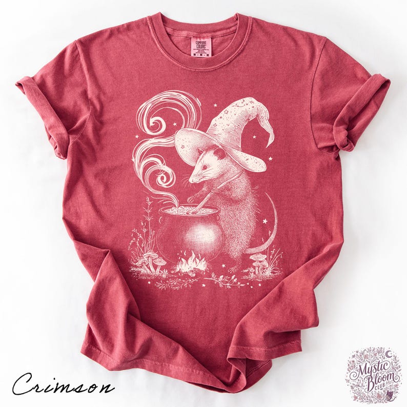 May include: A crimson short-sleeve t-shirt featuring a white graphic illustration of an opossum wearing a wizard hat and stirring a cauldron over a small fire. The design includes swirling smoke, stars, and mushrooms, with the text Comfort Colors.