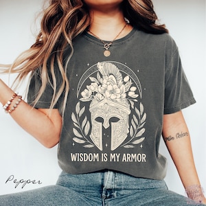 May include: A charcoal gray short-sleeve t-shirt featuring a cream-colored graphic of a Spartan helmet adorned with flowers and leaves. Above the helmet is a semi-circular arch, and below it is the text "WISDOM IS MY ARMOR" in a bold font.