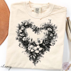 以下が含まれることがあります： アイボリー色のTシャツで、様々な花や葉で構成されたハート型の白黒グラフィックプリントが施されています。Tシャツにはクルーネックがあり、襟の内側部分にはComfort Colorsのブランドラベルがプリントされています。