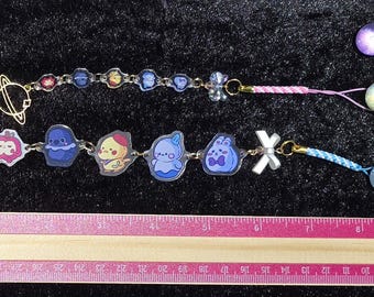 May include: Two charm bracelets with cartoon character charms and gold-colored planet charms. One bracelet has a pink and white cord, the other has a blue cord. A ruler is at the bottom of the image.