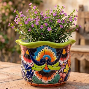 May include: A ceramic flower pot with a scalloped lime green rim and a vibrant, multi-coloured hand-painted floral pattern featuring orange, blue, red, and yellow hues. The pot contains a small green plant with numerous tiny purple flowers.