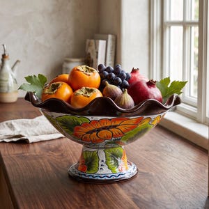 May include: A ceramic pedestal bowl with a ruffled rim, featuring hand-painted orange sunflowers and green leaves on a white background, filled with a variety of fresh fruit including persimmons, grapes, figs, and pomegranates.