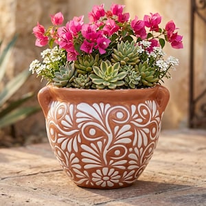 May include: A terracotta planter with two side handles and a white painted floral scroll pattern. The pot is filled with a vibrant arrangement of bright pink bougainvillea flowers, green succulent plants, and small white flowering sprigs.