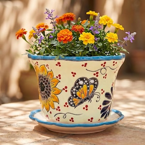 May include: A cream-colored ceramic flower pot with a matching saucer, featuring hand-painted yellow sunflowers, a black and orange butterfly, and small red dots. The pot is filled with a vibrant arrangement of orange, yellow, and purple flowers.