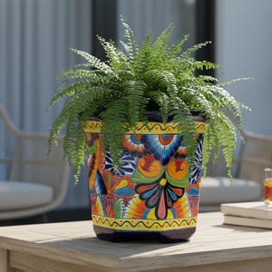 May include: A vibrant, hand-painted ceramic flower pot filled with a lush green fern. The pot features a colorful floral design with blue, yellow, orange, and green hues. It sits on a wooden table, creating a cheerful outdoor scene.