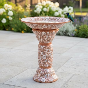 May include: A terracotta birdbath featuring a wide, shallow basin and a cylindrical pedestal base, both decorated with intricate white floral and swirling vine patterns. A small, iridescent blue and green hummingbird is perched on the rim.