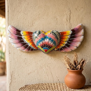 May include: A ceramic wall ornament shaped like a heart with outstretched wings. The piece features a vibrant, multi-colored hand-painted floral pattern in shades of pink, yellow, blue, green, and black, with a glossy finish on the surface.