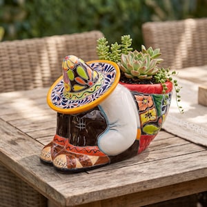 May include: A ceramic planter shaped like a person sitting with knees tucked, wearing a wide-brimmed sombrero with yellow, orange, green, and blue floral patterns. The planter holds green succulents and features hand-painted colourful designs.