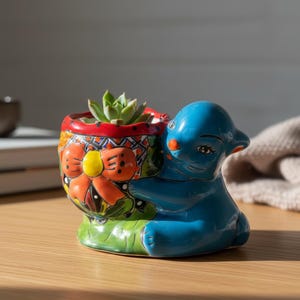 May include: A vibrant, hand-painted ceramic planter in the shape of a blue seal holding a colorful pot. The pot features a red rim, an orange bow, and a green base, with a succulent plant inside. A unique decorative item.