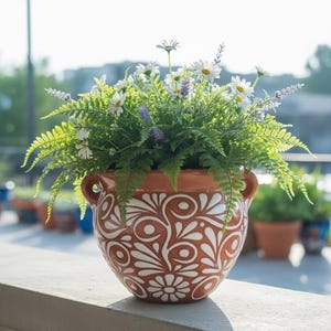 May include: A terracotta-colored ceramic flower pot with a white floral design, filled with green ferns, white daisies, and purple lavender. The pot has two small handles and sits on a concrete ledge.