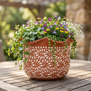 May include: A terracotta-colored ceramic planter with a scalloped rim and a repeating white floral pattern. The pot is filled with a variety of trailing green ivy, small green bead-like succulents, and clusters of tiny purple, yellow, and white flowers.