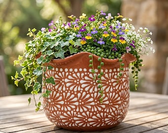 May include: A terracotta-colored ceramic planter with a scalloped rim and a repeating white floral pattern. The pot is filled with a variety of trailing green ivy, small green bead-like succulents, and clusters of tiny purple, yellow, and white flowers.