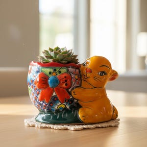 May include: A colorful ceramic planter with a succulent plant. The planter features a detailed design with a large red bow, floral patterns, and a yellow lion cub figure. The planter sits on a small, round, cream-colored doily.