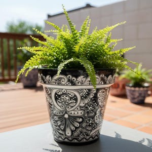 May include: A black and white decorative ceramic flower pot with a fern plant. The pot has a detailed floral design. The fern has bright green leaves. The pot is sitting on a gray surface.