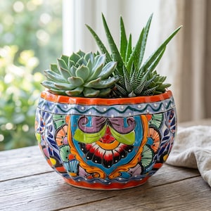 May include: A round ceramic planter featuring a vibrant, multi-colored hand-painted floral pattern in shades of orange, blue, green, and purple. The planter contains a cluster of green succulents, including an aloe plant and a rosette-shaped succulent.