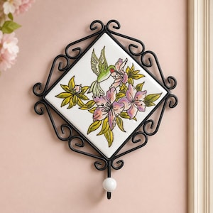 May include: A diamond-shaped ceramic wall hook featuring a painted illustration of a green hummingbird hovering near pink and yellow flowers. The tile is framed by a black metal scrollwork border and includes a single white knob at the bottom.