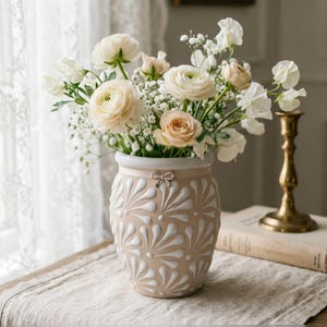 May include: A beige ceramic vase with a white rim and a repeating white fan-shaped embossed pattern. The vase features a small metallic bow detail at the neck and is filled with a bouquet of white and pale peach ranunculus and baby's breath flowers.