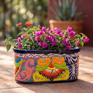 May include: An oval ceramic planter featuring hand-painted, vibrant floral and scroll patterns in shades of yellow, orange, green, pink, and blue. The planter is filled with bright magenta bougainvillea flowers and small orange lantana blooms.
