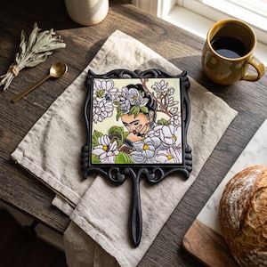 May include: A square ceramic tile featuring a colorful illustration of a person with dark hair adorned with white flowers, surrounded by botanical elements. The tile is set within a black, ornate, cast-iron frame with a handle extending from the bottom.
