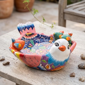 May include: A hand-painted ceramic bird-shaped bowl featuring a white head with an orange beak and black eyes. The bowl is decorated with vibrant floral patterns in shades of pink, blue, purple, and yellow, with small bird figures attached to the rim.