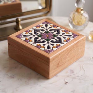 May include: A square wooden box featuring a decorative ceramic tile inlay on the lid. The tile displays a symmetrical floral pattern in shades of purple, dark blue, green, and orange against a white background. The box has a natural wood grain finish.