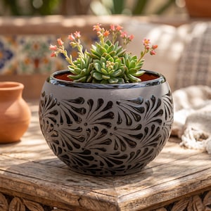May include: A round ceramic planter with a matte grey finish and a glossy black hand-painted floral pattern. The planter features a wide opening with a black rim and contains a green succulent plant with small, delicate orange-pink flowers.