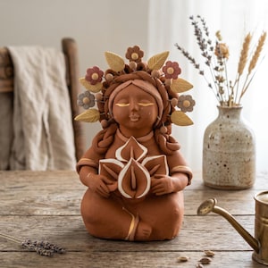 May include: A terracotta-colored clay figurine of a person with closed eyes, adorned with a crown of sculpted flowers and leaves. The figure holds a cluster of white-tipped calla lily-like flowers and features gold painted accents on the eyes.