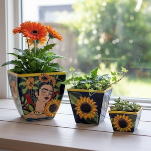 May include: A trio of square ceramic plant pots. The largest pot displays a woman's portrait with sunflowers. The other two pots feature sunflower designs. The pots are dark blue with yellow trim and contain plants and flowers.