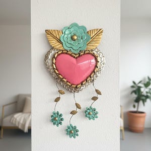 May include: A decorative metal heart wall hanging. The heart is pink with a gold border, surrounded by a silver filigree. It features a turquoise flower and leaves at the top, with turquoise flowers hanging below. The piece is mounted on a white wall.