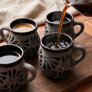 May include: Three round ceramic mugs with a matte grey finish and black painted floral patterns. Each mug features a dark black rim and handle. Dark coffee is being poured from a glass carafe into one of the mugs, creating ripples in the liquid.