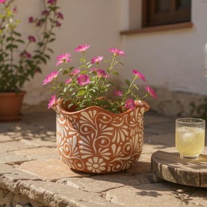 May include: A terracotta-colored ceramic flower pot with a white floral design, filled with vibrant pink flowers. The pot sits on a stone surface, with a glass of iced beverage nearby. The pot has a scalloped rim.