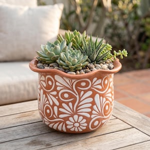 May include: A terracotta planter with a scalloped rim and white floral scrollwork patterns. The pot is filled with a variety of green succulents and small grey pebbles. The planter has a rounded body and a smooth, glazed finish on the exterior.