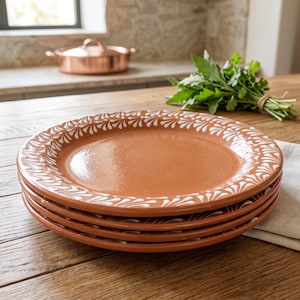 May include: A stack of four round terracotta plates featuring a smooth, glazed finish and a decorative white painted pattern along the rim. The plates are crafted from natural clay, showcasing a warm reddish-brown color and a consistent design.