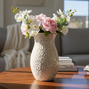 May include: A cream-colored ceramic vase with a detailed floral pattern holds a bouquet of white orchids and pink peonies. The vase sits on a wooden table, creating a warm and inviting aesthetic. The flowers add a touch of elegance to the room.