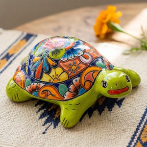 May include: A ceramic turtle figurine featuring a bright green head and limbs, with a shell decorated in a vibrant, multi-colored floral pattern including shades of orange, yellow, blue, and red. The turtle has a smooth, glossy finish.