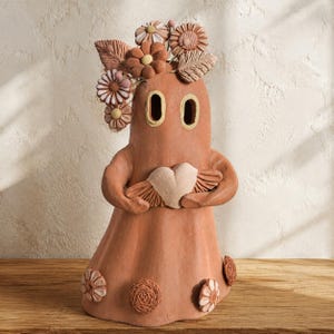 May include: A terracotta-colored ceramic ghost figurine featuring two oval eye cutouts, holding a light-colored heart with wings. The figure is adorned with sculpted floral and leaf accents on its head and base, showcasing a matte, earthy finish.