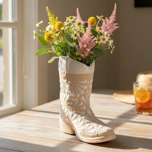 May include: A cream-colored ceramic cowboy boot vase filled with a vibrant bouquet of yellow, white, and pink flowers. The boot features embossed floral patterns and a two-tone design. The vase is displayed on a wooden surface.